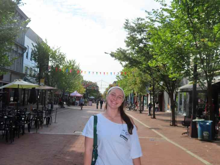 A young woman in a white shirt and denim shorts stands on a brick street lined with trees and colorful bunting.
