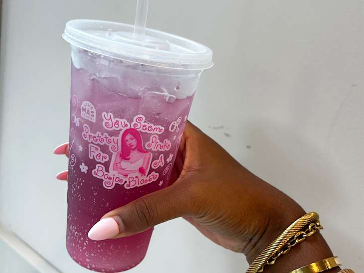 A hand holding a clear cup with a pink drink and ice, featuring decorative text and a straw, against a light background.