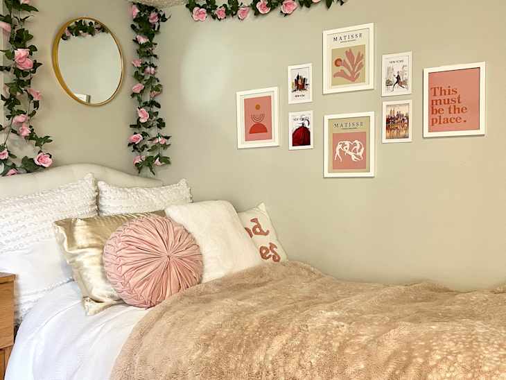 Dorm room with pale green walls, bed with pink and white bedding and throw pillows, faux pink roses and leaves hanging down walls, over bed, gallery wall by bed of pink and white art