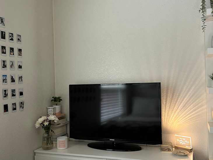 corner of a white dresser with TV, vase of white flowers, stack of books with plant on top, jar candle, some polaroids on the wall