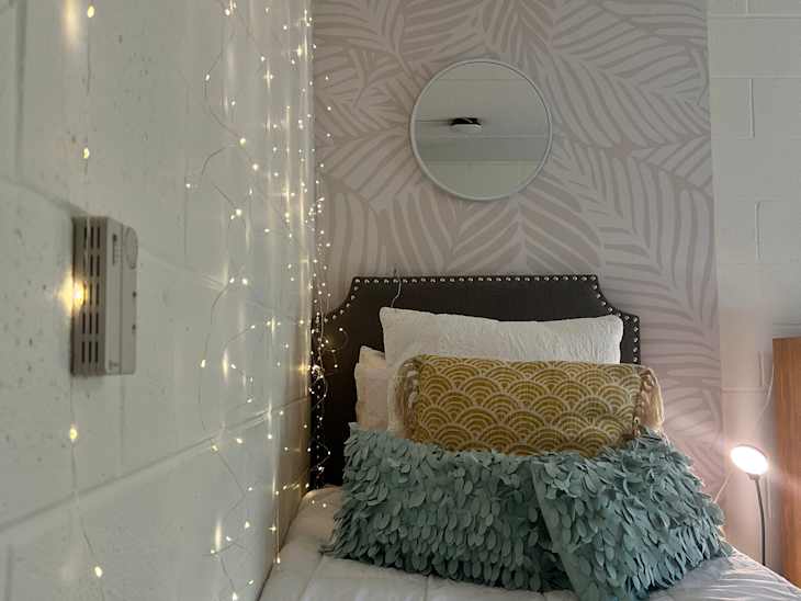 raised bed with white bedding and string lights hanging down wall, round mirror above bed, leaf wallpaper on wall behind bed