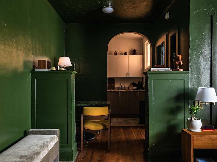 Green walls and ceiling, small half pillar room dividers, gray velvet bench, yellow mid century modern chair, small wood night stand, arched entrance to kitchen