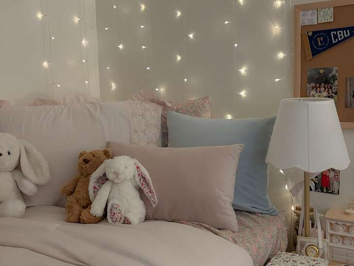 Cozy bedroom with fairy lights, plush toys, pastel bedding, and a bedside lamp on a wooden nightstand.