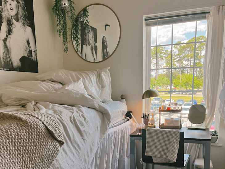Cozy bedroom with a lofted bed, white bedding, a desk with a lamp, and a large window overlooking trees and parked cars.