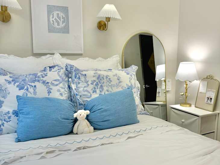 Cozy bedroom with a white bedspread, blue floral pillows, a stuffed lamb, a gold lamp, and a mirror.