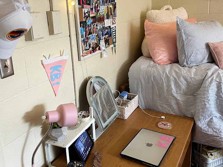 Dorm room setup featuring a bed with striped bedding, decorative pillows, a pink lamp, and a laptop on a wooden desk.