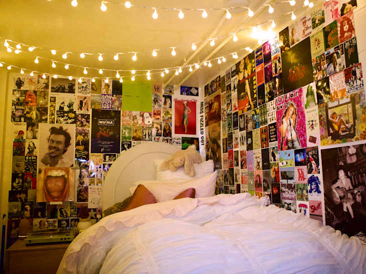 Cozy bedroom with a white bedspread, decorated walls covered in posters, and string lights overhead.