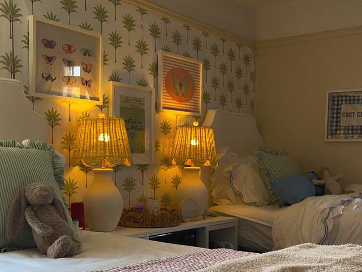 Cozy bedroom with palm-patterned wallpaper, two lamps, plush toys, and layered bedding in soft colors.