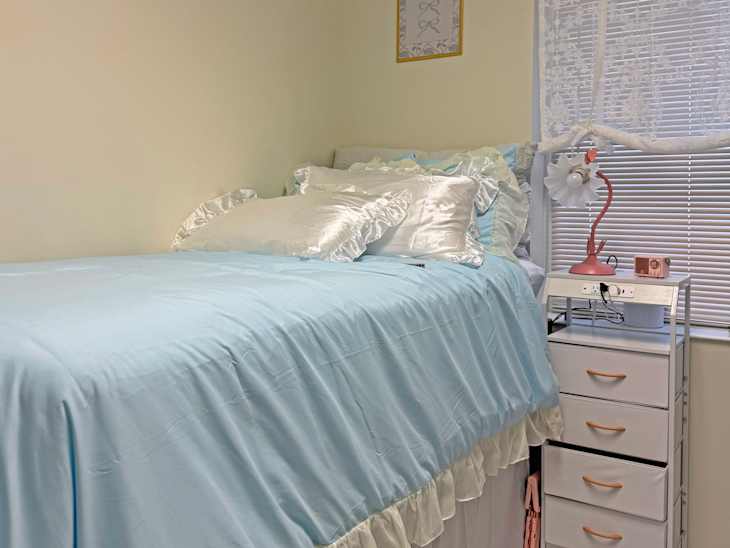Cozy bedroom with a light blue bedspread, decorative pillows, a pink lamp, and a small storage unit beside the bed.