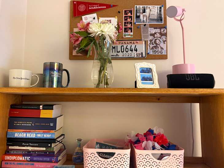 Desk shelf with two mugs, a vase of flowers, a pink lamp, JBL speaker, framed photo, and stacked books.