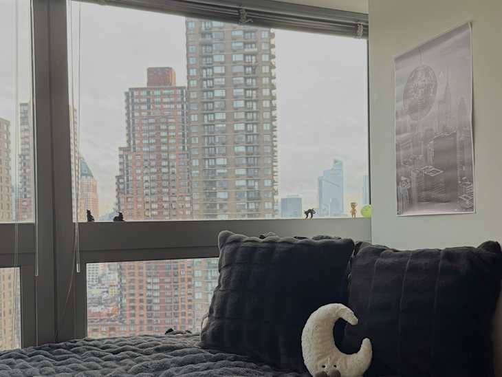 Cozy bedroom corner with a black plush bedspread, two large pillows, and a decorative plush moon, overlooking city skyscrapers.