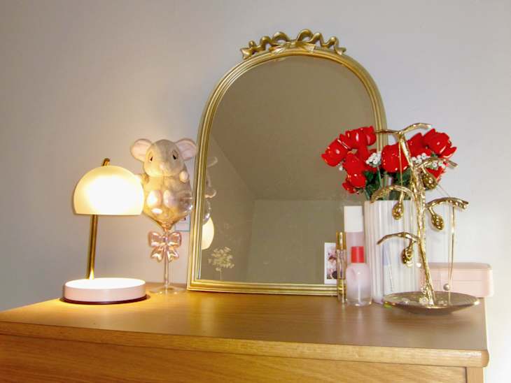 Dresser with a gold-framed mirror, a lamp, a plush elephant, red roses, and various beauty products.