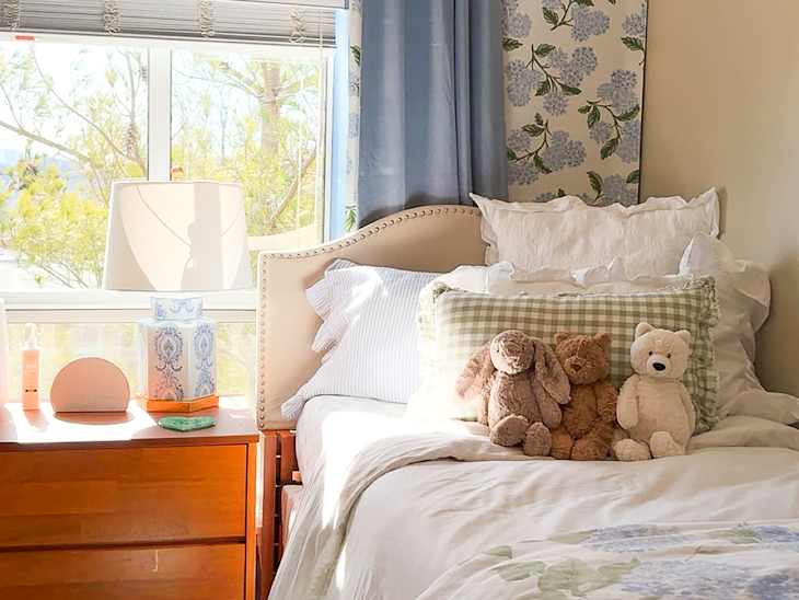 Cozy bedroom with floral wallpaper, a bed adorned with plush toys, and a wooden nightstand holding decor items.