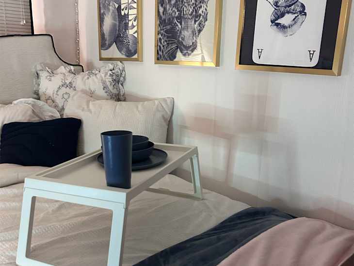 Cozy bedroom scene featuring a bed with white bedding, decorative pillows, and a tray with blue dishes. Wall art includes framed prints.