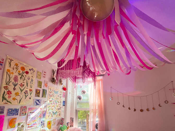 Colorful ceiling decorations with pink and white streamers, floral wall art, and a window with sheer curtains.
