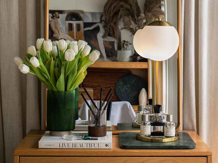 dorm room after installing diy curtain wall. Neutral curtain wall behind wood nightstand with arched mirror, books, white tulips, perfume, small lamp.