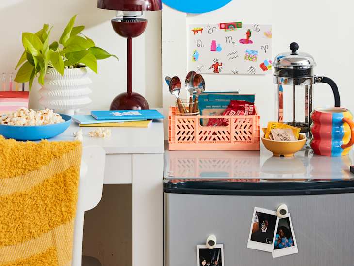 closeup shot of desk that has a red lamp, white planter, supplies and a blue bowl of popcorn. next to the desk is a mini fridge with a mug, coffee press, and a pink basket with utensils coffee assortments on it