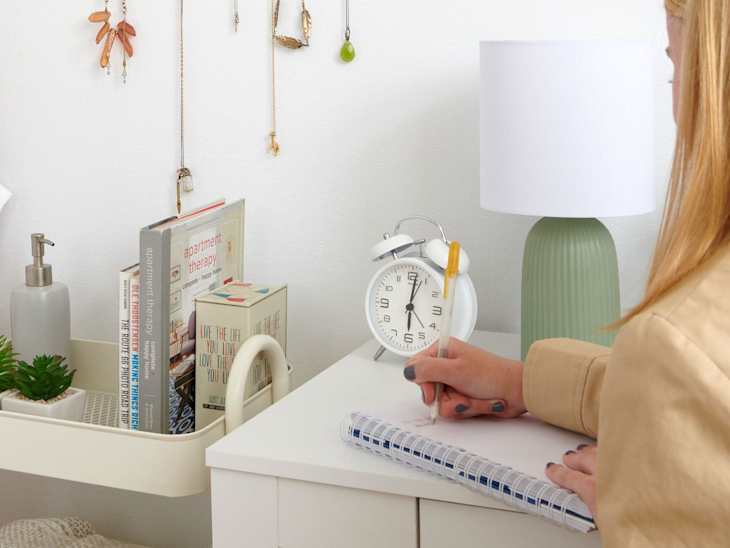 angled shot of wall organizer with jewelry hanging off of it and sunglasses at top and a person writing on a notebook at desk