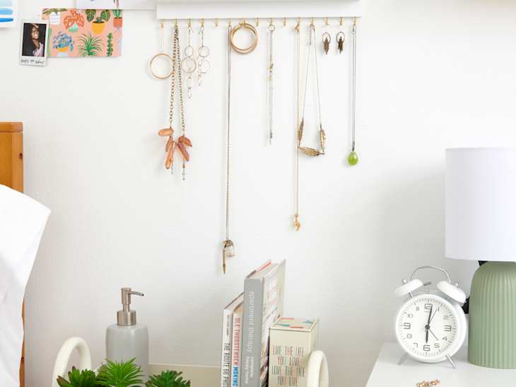 angled close shot of wall organizer with jewelry hanging off of it and sunglasses at top and a desk with a clock, lamp, and jewelry