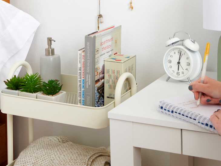 person writing on a notebook at desk next to white rolling card with books, plants. bed with white pillow to the far left
