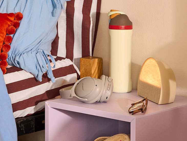 A bedside table with a lavender finish, featuring headphones, a water bottle, a wooden block, and a lamp.