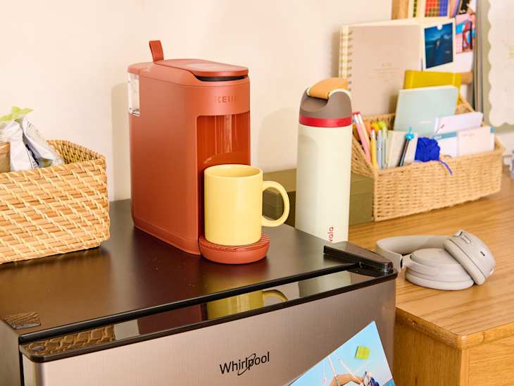 Keurig coffee maker in terracotta color with a yellow mug, a water bottle, and a basket of snacks on a wooden table.