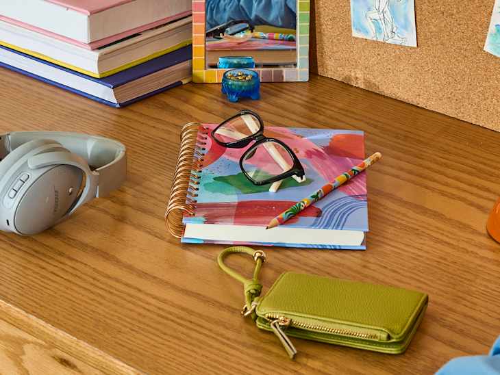 Colorful notebook with glasses, a patterned pencil, headphones, and a green wallet on a wooden desk.