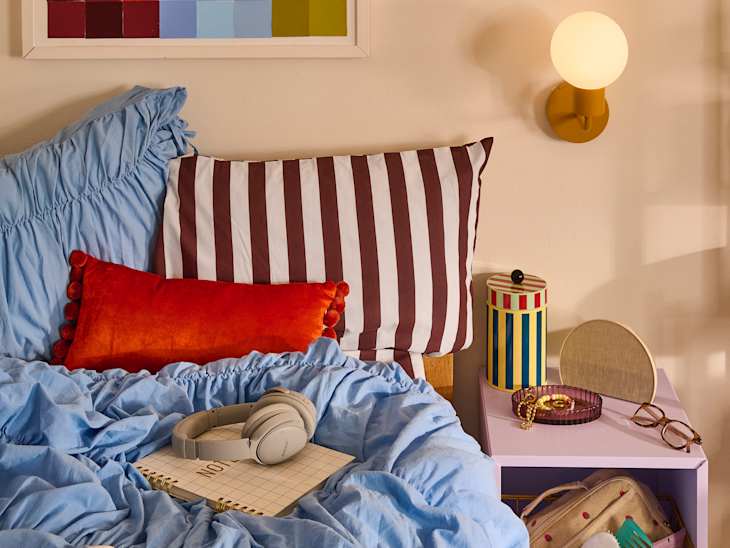 Cozy bed with blue ruffled bedding, striped and orange pillows, headphones, and colorful decor on a nightstand.