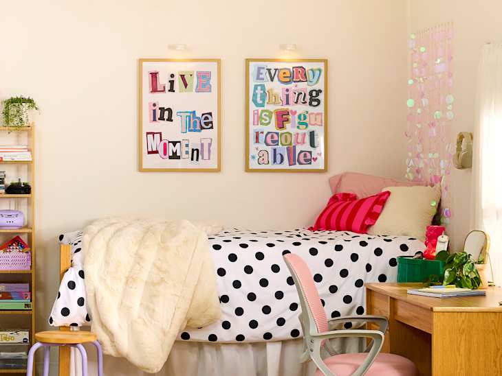 Cozy bedroom with a polka dot bedspread, pink pillows, framed motivational art, and a wooden desk with a green plant.