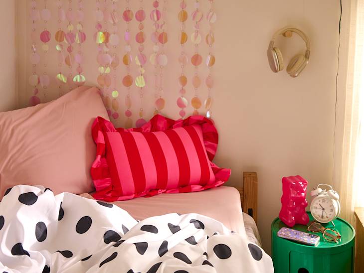 Cozy bedroom corner with a pink bed, striped pillow, polka dot blanket, green nightstand, alarm clock, and headphones.