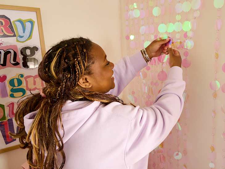 A person with braided hair hangs colorful circular decorations on a wall, wearing a light purple hoodie.
