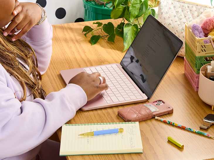 A person typing on a pink laptop at a wooden desk with a notepad, pen, phone case, and colorful stationery.
