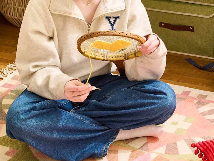 Person sitting cross-legged on a colorful rug, using a wooden embroidery hoop with yellow yarn.