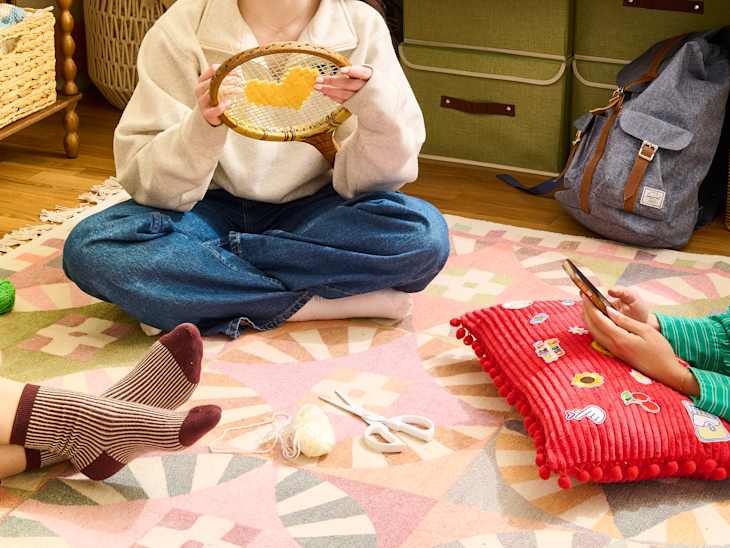 A cozy indoor scene with three people crafting on a colorful rug, holding yarn, scissors, and a phone.