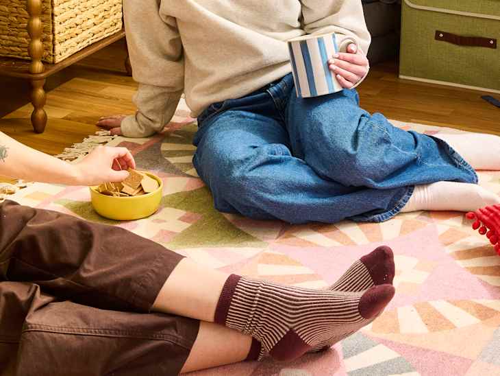 Two people sitting on a colorful rug, one holding a striped mug and the other reaching for snacks in a yellow bowl.