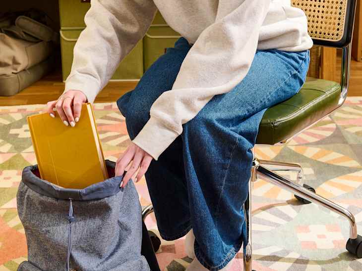 A person in a gray sweatshirt and blue jeans is placing a yellow notebook into a gray backpack while sitting on a green chair.