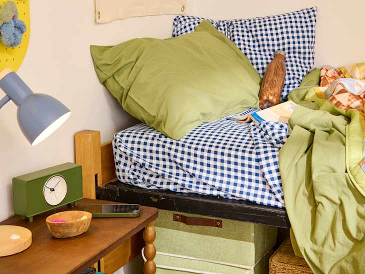 Cozy bed with green and blue checkered bedding, a wooden nightstand, a green clock, and a lamp.