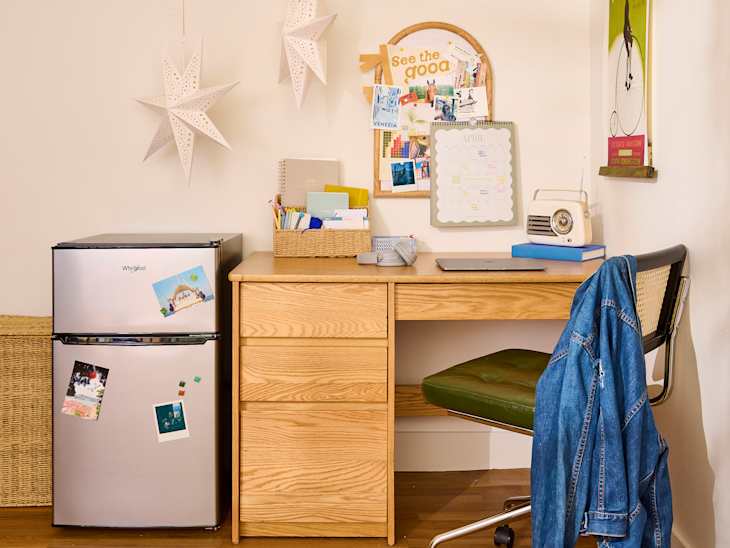 Compact workspace featuring a wooden desk, green chair, mini fridge, and decorative wall items.
