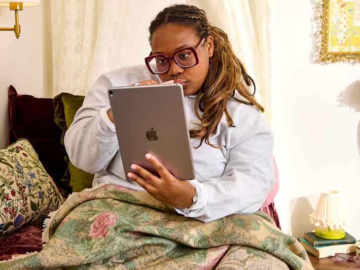 A person in a gray sweatshirt sits on a couch, using a tablet while wrapped in a floral blanket, surrounded by decorative pillows.