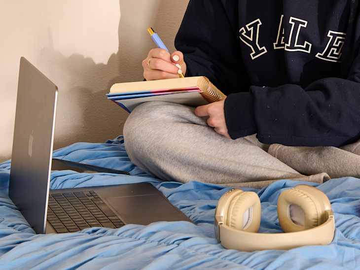 A person wearing a Yale sweatshirt sits on a blue bedspread, writing in a notebook with a blue pen, next to a laptop and headphones.