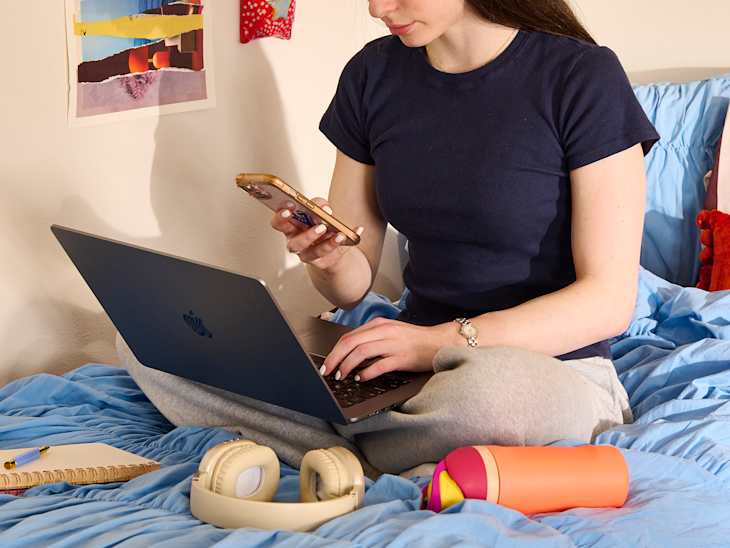 A person sitting on a bed with a blue comforter, using a laptop and holding a phone, with headphones and a colorful container nearby.