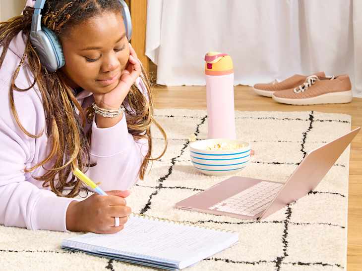 A young person with headphones lies on a rug, writing in a notebook, with a laptop, popcorn bowl, and water bottle nearby.
