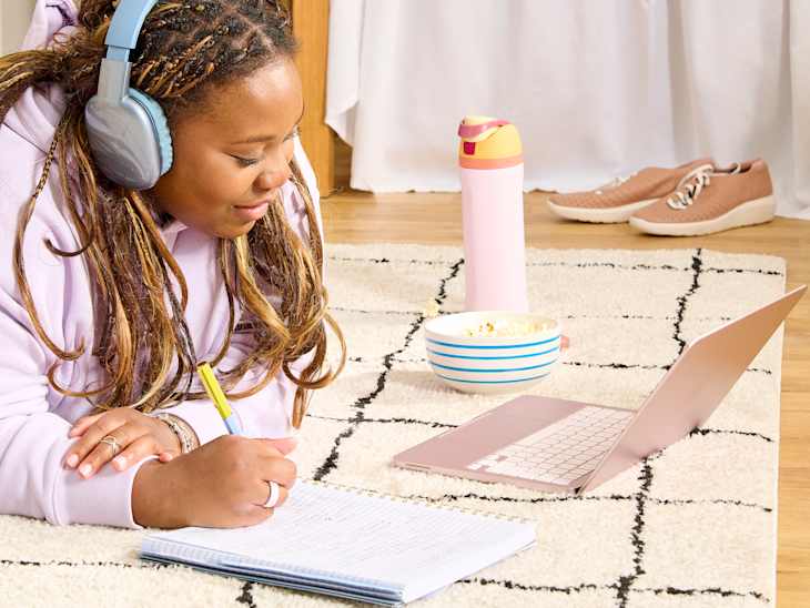 A young woman with headphones writes in a notebook on a rug, with a laptop, popcorn bowl, and water bottle nearby.