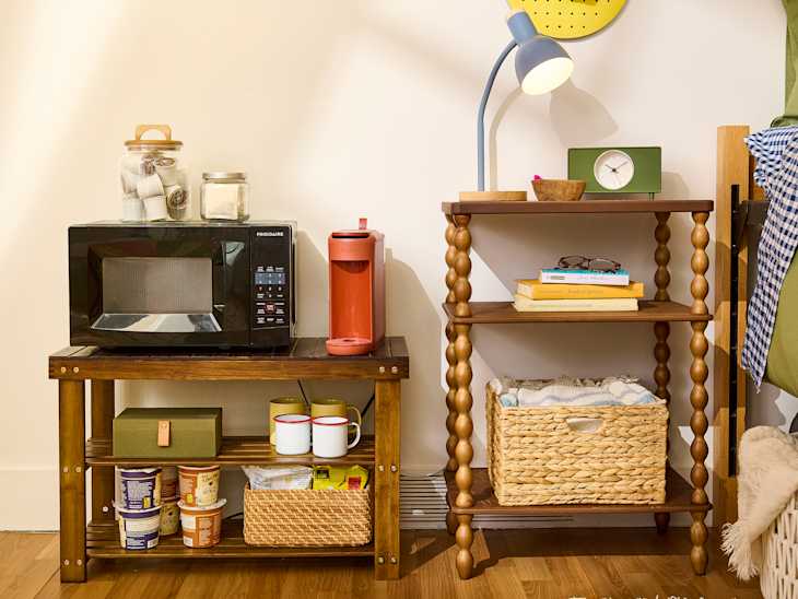 Microwave and coffee maker on a wooden shelf, with books, glasses, and a woven basket on a nearby table.