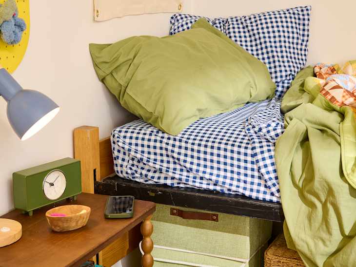 Cozy bed with blue and white checkered bedding, green pillows, a green clock, and a wooden nightstand.