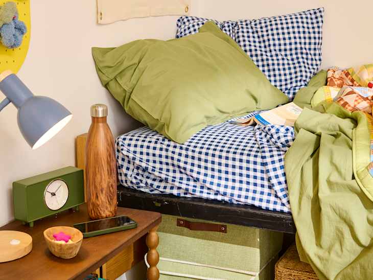 Cozy bed with green and blue checkered bedding, wooden nightstand, lamp, water bottle, and a smartphone.