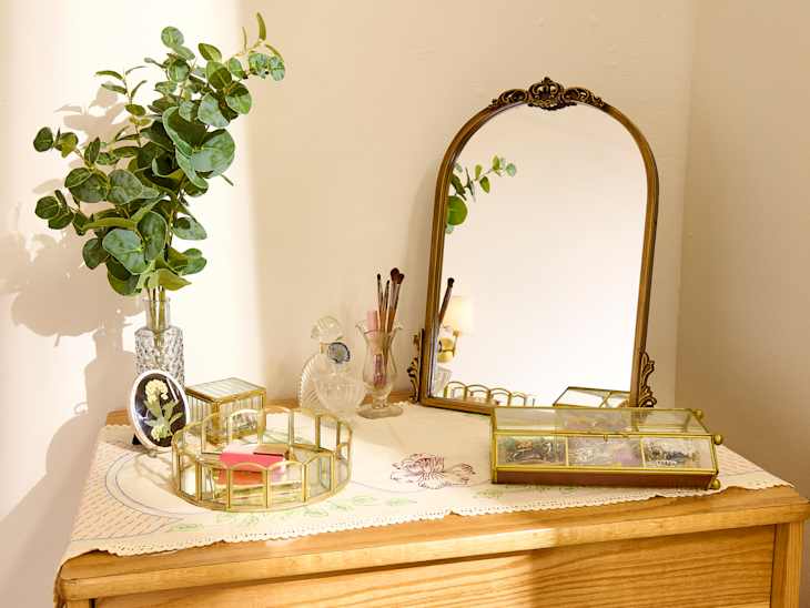A wooden dresser topped with a vintage mirror, decorative trays, a vase of eucalyptus, and makeup brushes.