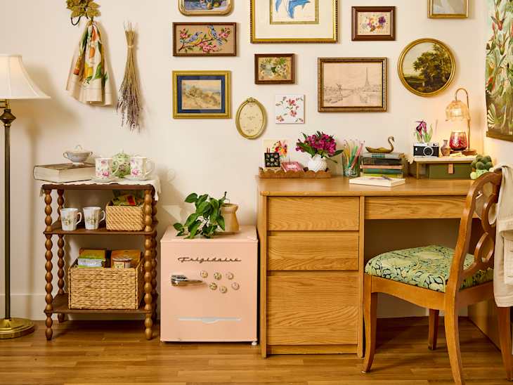 Cozy workspace with a wooden desk, pink fridge, decorative wall art, and a plant on a side table.