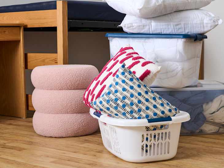 Bedding and decorative pillows stacked in a laundry basket next to a pink stool and wooden desk.