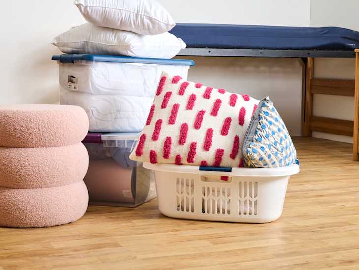Bedding and decorative pillows stacked in a laundry basket, with a pink pouf and a bed frame in the background.